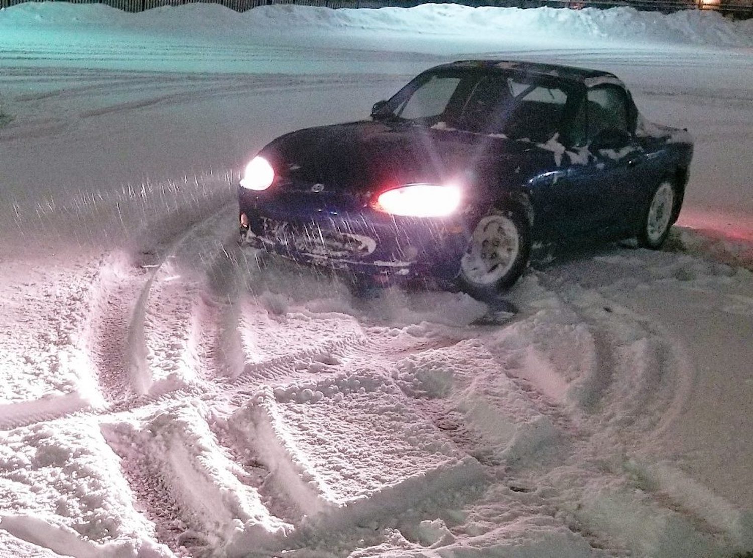 A Mazda Miata roadster parked in a circle of tire tracks left in a snow-covered parking lot.