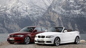 A red 2012 E82 BMW 135i Coupe next to a white 2012 E88 BMW 135i Convertible by a mountain lake
