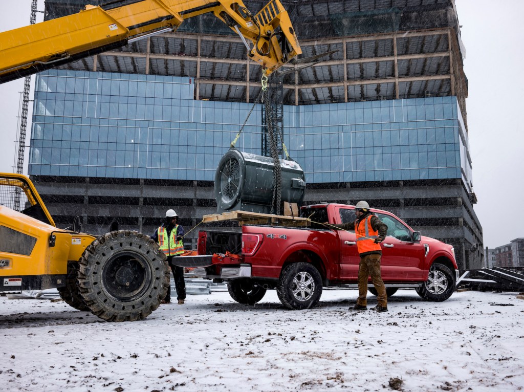 Construction workers loading heavy equipment in the bed of a red Ford F-150 in front of a half-finished building.