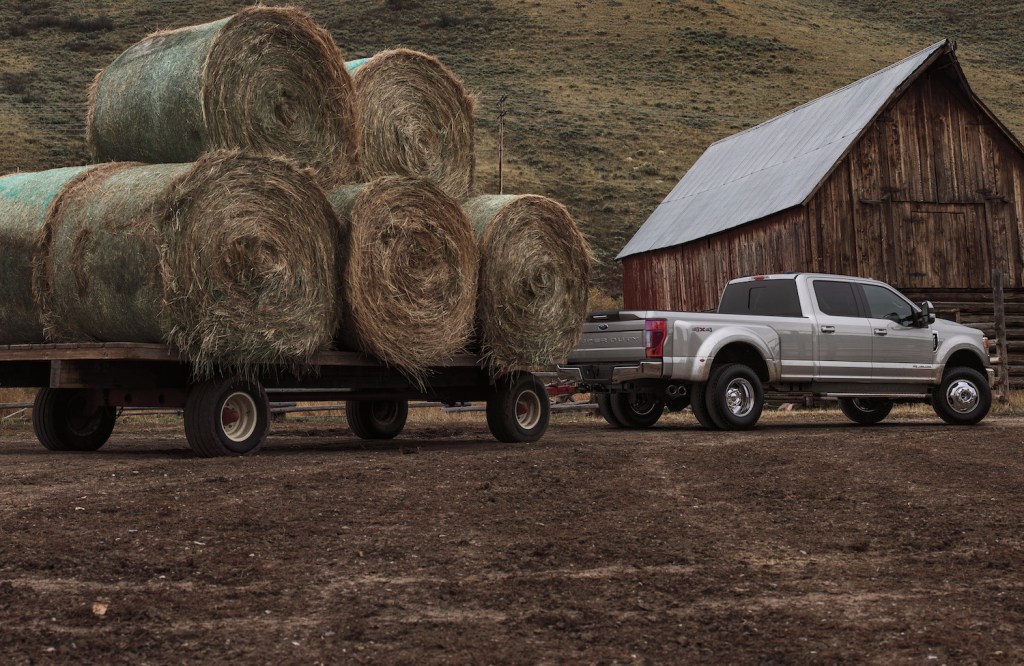 Silver Ford F-Series F 250 Super Duty truck towing a trailer loaded with hay bales in front of a barn.