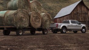 Silver Ford F-Series F 250 Super Duty truck towing a trailer loaded with hay bales in front of a barn.