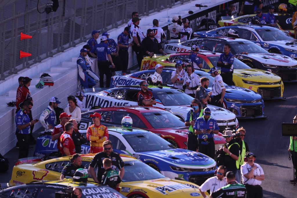 Drivers and team members stand by a row of NASCAR race cars before a race.