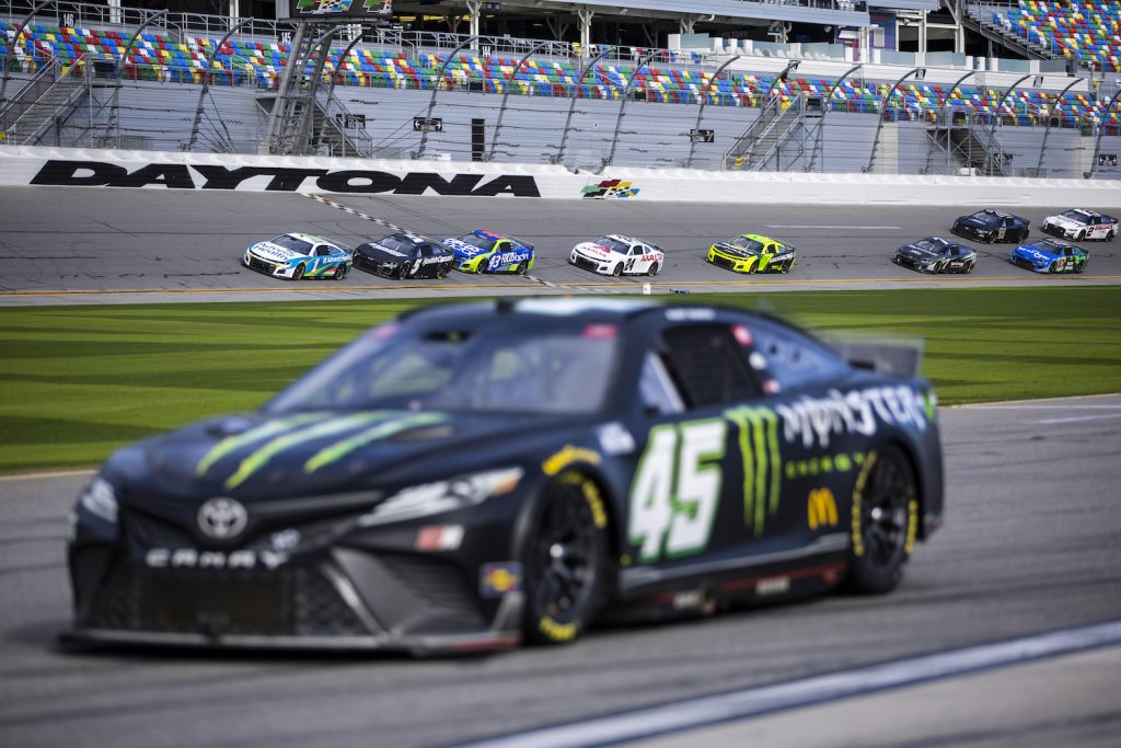 A NASCAR race car parked in pit lane at Daytona with a row of other cars racing by in the background.