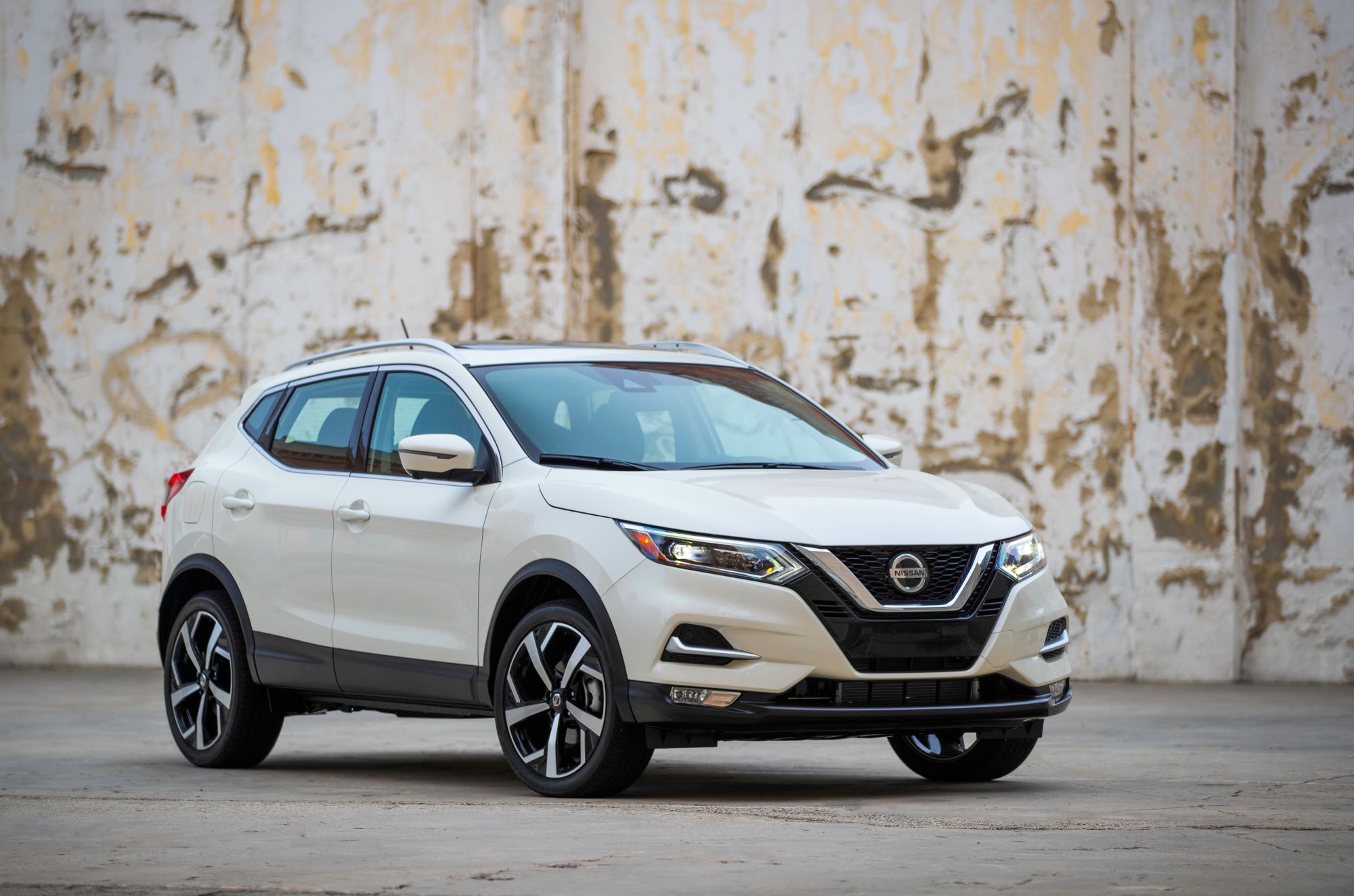 A white 2022 Nissan Rogue Sport on blacktop in front of a distressed concrete white painted wall.