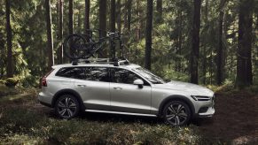 A profile view of a silver Volvo V60 Cross Country parked in the woods with a bike rack and bikes on the roof.