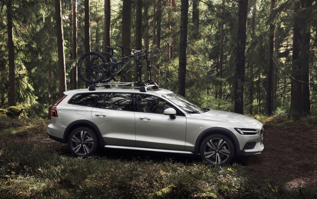 A profile view of a silver Volvo V60 Cross Country parked in the woods with a bike rack and bikes on the roof.