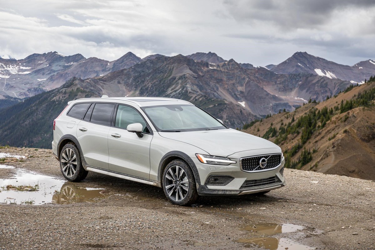 A 3/4 front view of a silver Volvo V60 Cross Country parked on gravel next to a puddle with mountains in the background.
