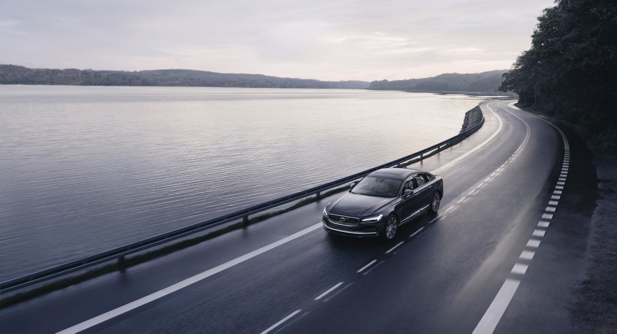 A 3/4 front and overhead view of a gray Volvo S90 Recharge driving on a road with a lake in the background
