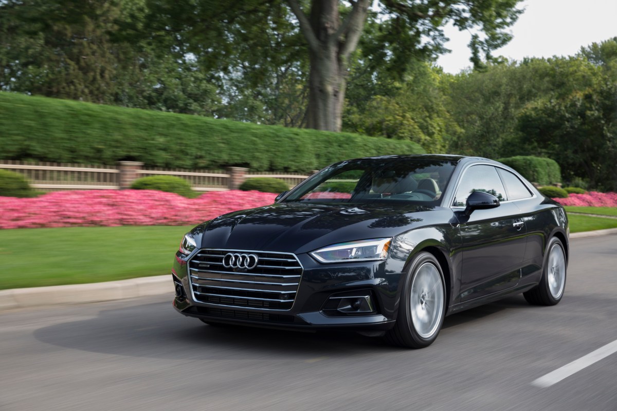 A 3/4 front view of a black 2020 Audi A5 Coupe driving on a road with flowers in the background.