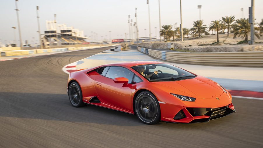 A 3/4 front view of an orange Lamborghini Huracan EVO cornering on a race track. Grand stands and palm trees are visible in the background.