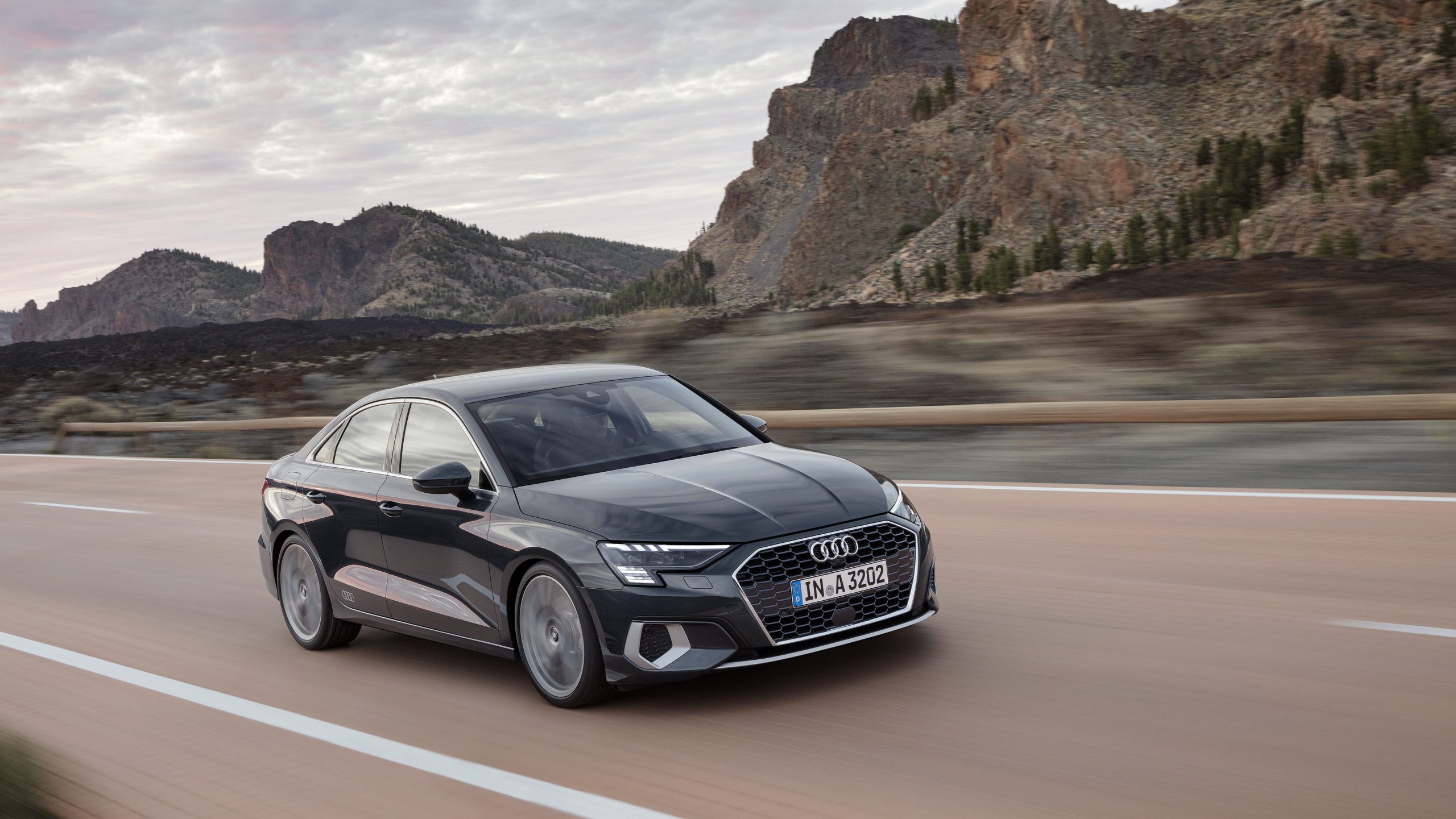 A 3/4 front view of a dark gray 2022 Audi A3 sedan driving on a road with mountains in the background.