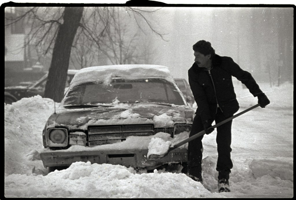 This is a man on a city street shoveling out his car after a snowstorm.