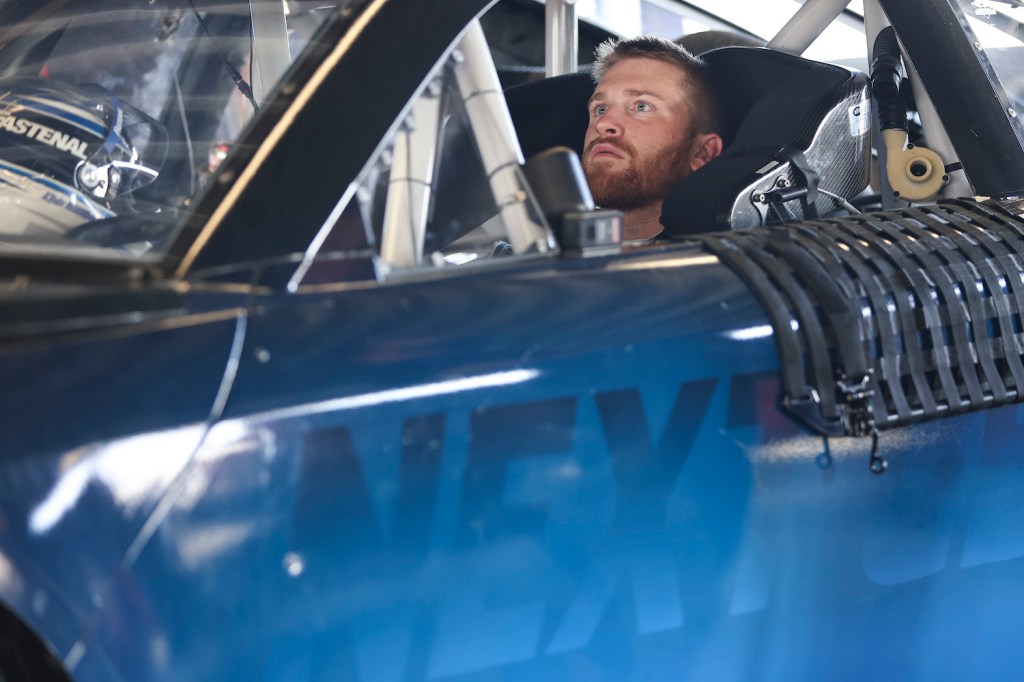 Driver Chris Buescher sits in a NASCAR car staring up at the rearview mirror or rearview camera screen.