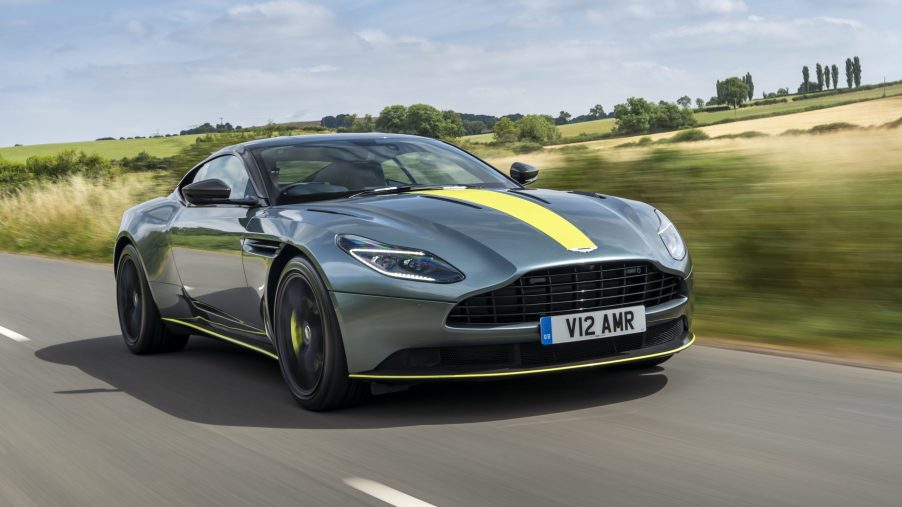 A 3/4 front view of a gray Aston Martin DB11 driving on a road with fields in the background.