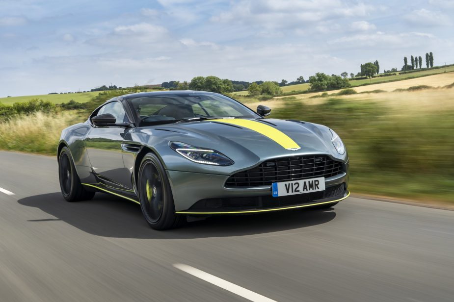 A 3/4 front view of a gray Aston Martin DB11 driving on a road with fields in the background.