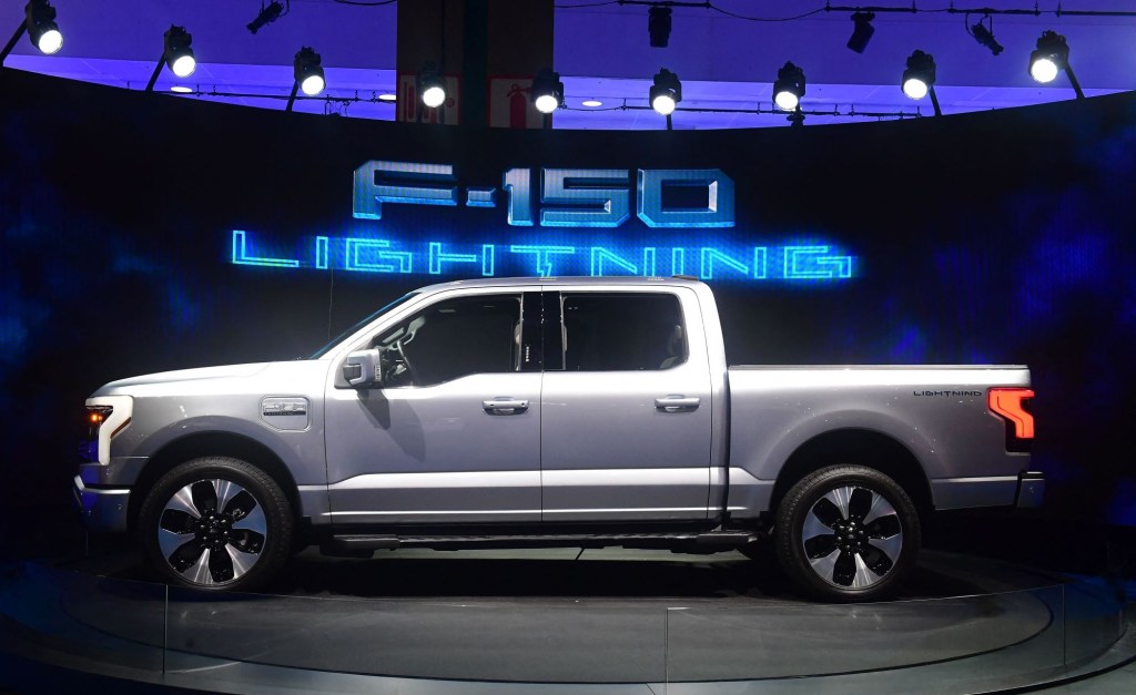 A silver Ford F-150 Lightning in front of a black background with F-150 Lightning written in blue lights in the background.