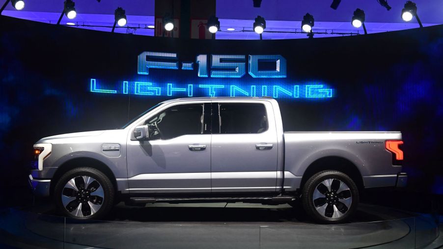 A silver Ford F-150 Lightning in front of a black background with F-150 Lightning written in blue lights in the background.