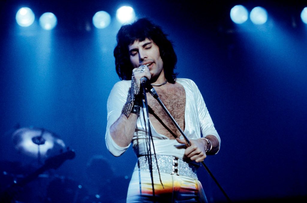 Freddie Mercury iš Queen scenoje dainuoja baltais marškiniais su atvira krūtine ir baltomis kelnėmis su sidabru blizgančiu diržu su juodomis sagomis su lemputėmis fone.