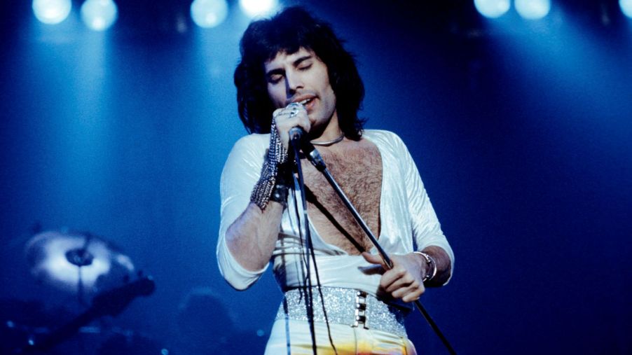 Freddie Mercury from Queen singing on stage in a white open-chest shirt and white pants with a silver glittery belt with black buttons with lights in the background.