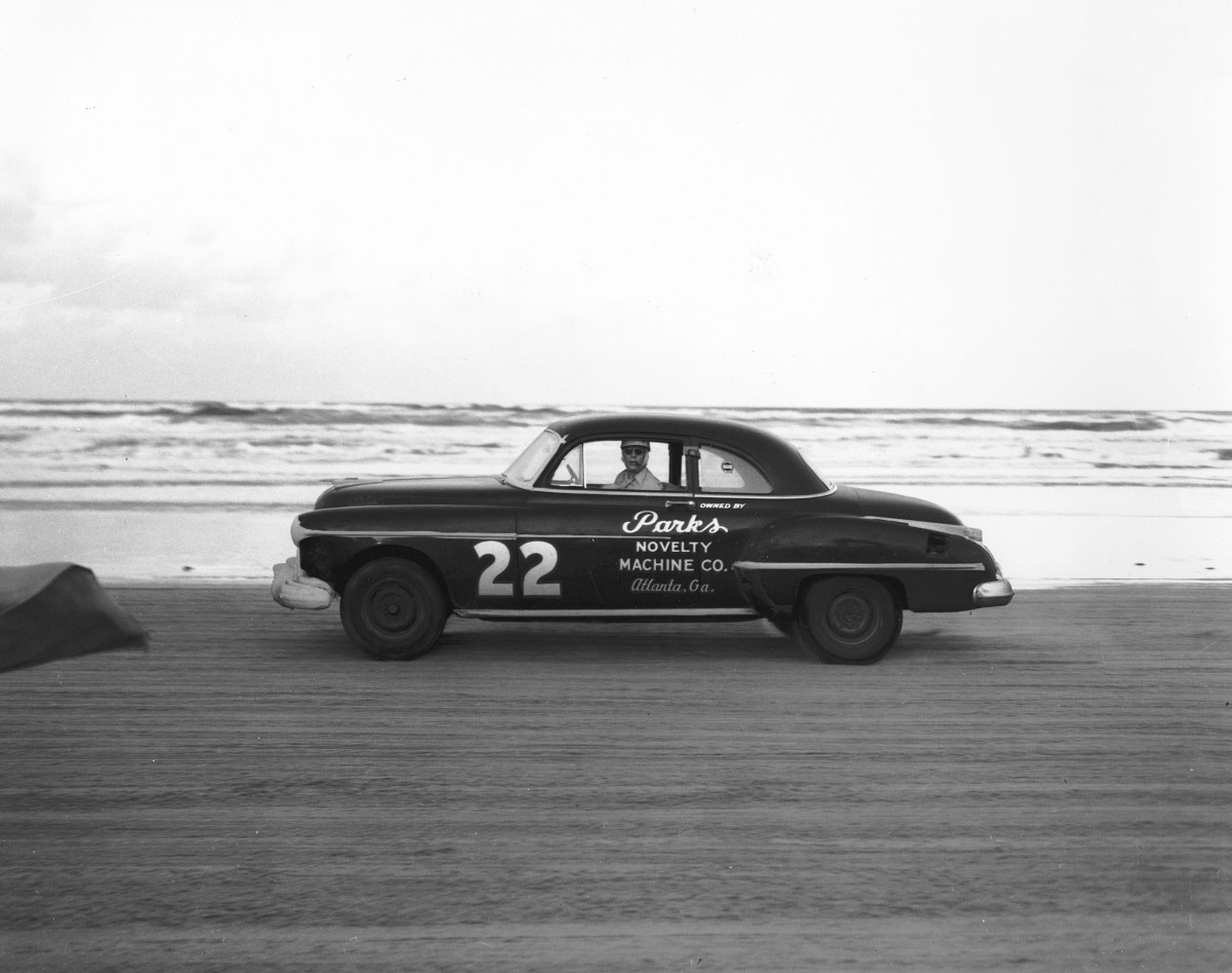 A black and white image of NASCAR driver Red Byron behind the wheel of a 1950 Oldsmobile 88 coupe driving on the original Daytona Beach racecourse.