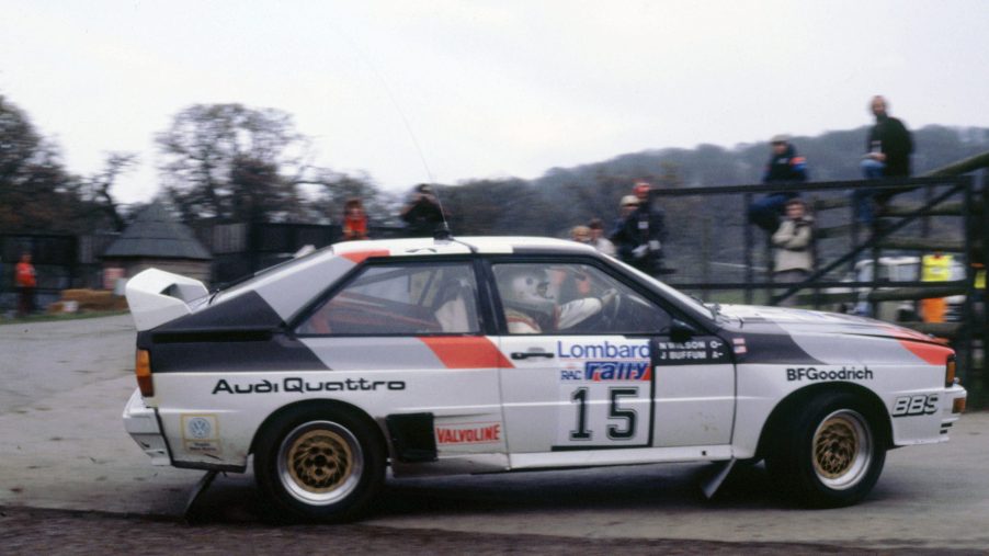 A profile view of a 1983 Audi Quattro rally car during a World Rally Championship race. The car is cornering hard with fans sitting in the background.