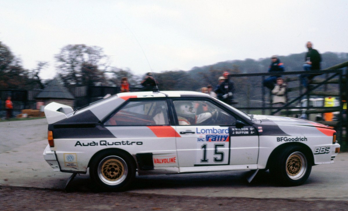 A profile view of a 1983 Audi Quattro rally car during a World Rally Championship race. The car is cornering hard with fans sitting in the background.