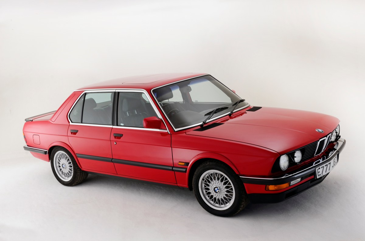A 3/4 profile view of a red 1987 BMW M5 sedan parked in a white studio background.
