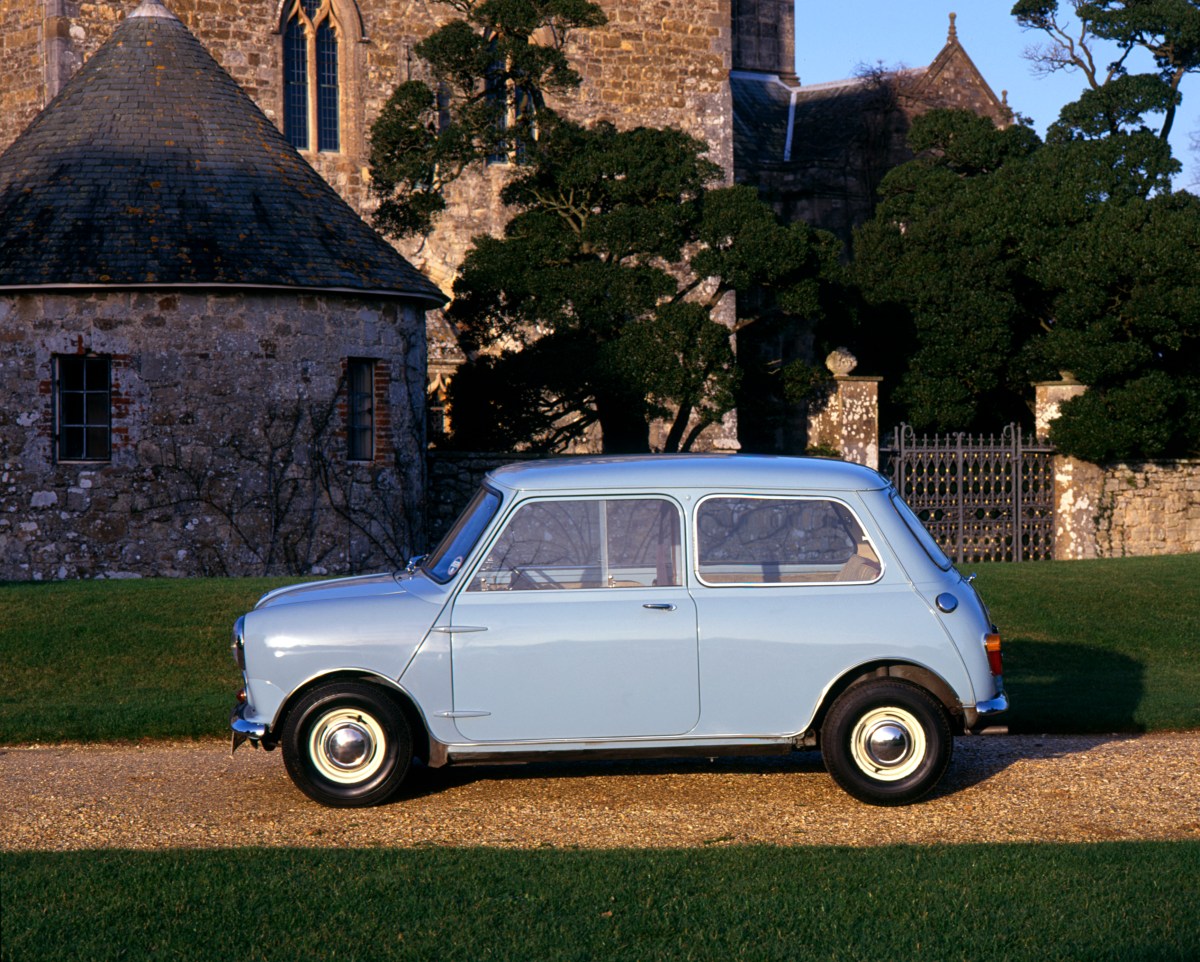 A profile view of a light blue 1959 Austin Mini Seven parked on a gravel road in front of a stone building.