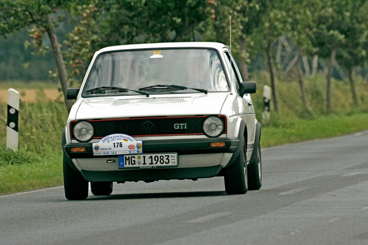 A front view of a white 1982 Volkswagen Golf GTI driving on a road with trees in the background.