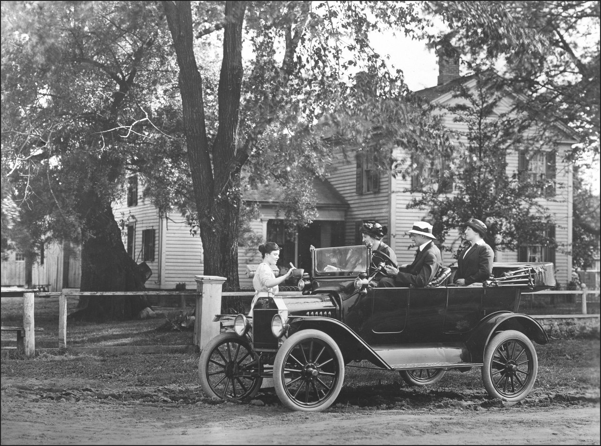 A black and white image from 1908 showing a Ford Model T parked on a dirt road in front of a fence with a driver and two passengers.