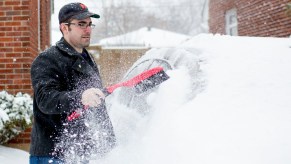 A man in a baseball cap stands in front of a brick house and uses a tool to brush the snow off a car.