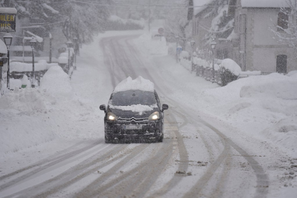 Black car driving down a snowy road in a blizzard, its roof unsafely piled with snow.
