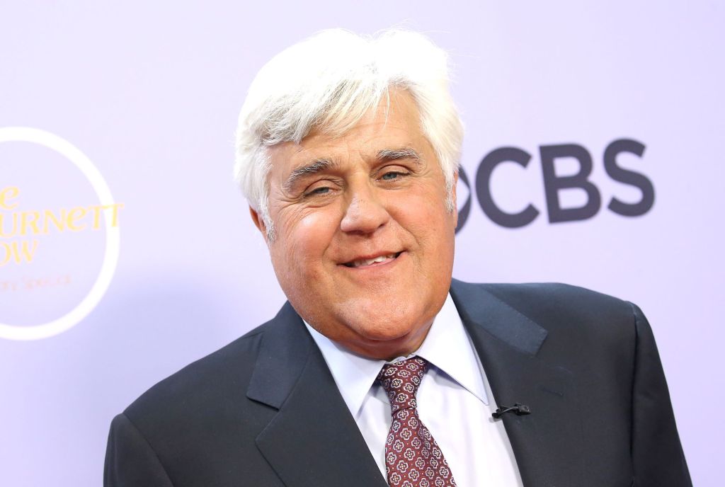 Jay Leno standing in front of a light blue background dressed in a black suit, white shirt, and a light red tie with dark red diamond outlines on it.