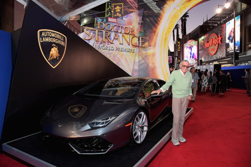 Stan Lee next to a Silver Lamborghini Huracán coupe standing on a red carpet.
