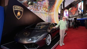 Stan Lee next to a Silver Lamborghini Huracán coupe standing on a red carpet.