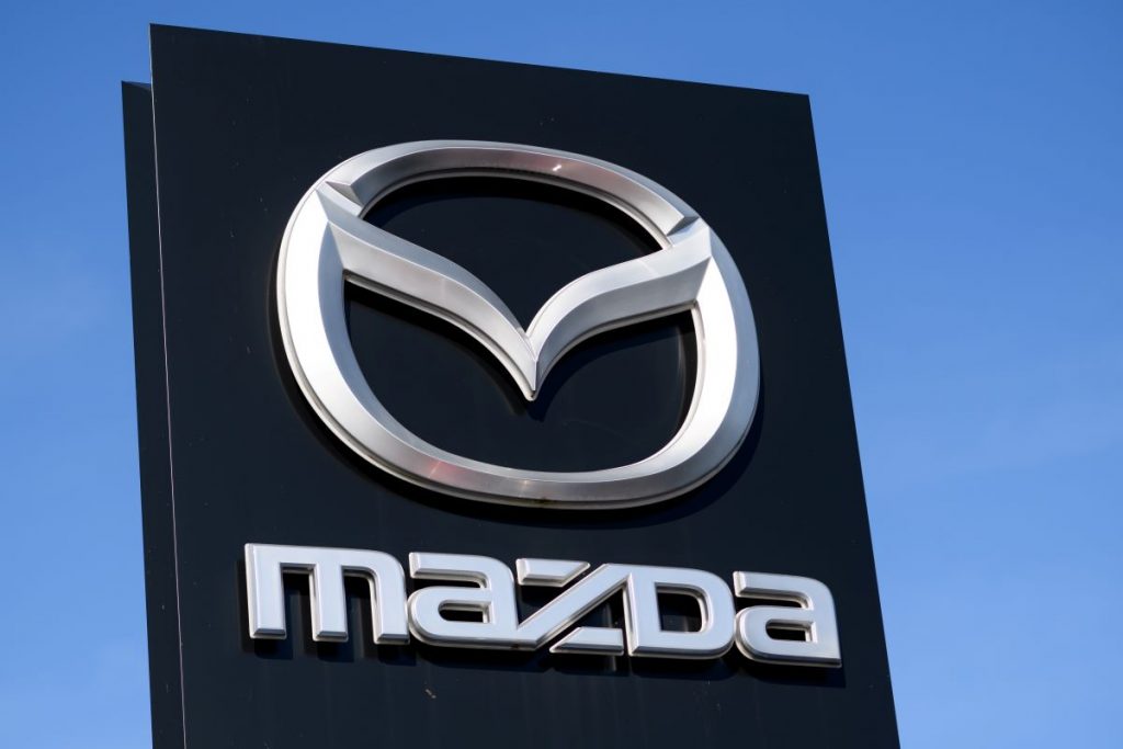 A black Mazda sign in front of a blue clear sky with the Mazda logo in silver on it. 