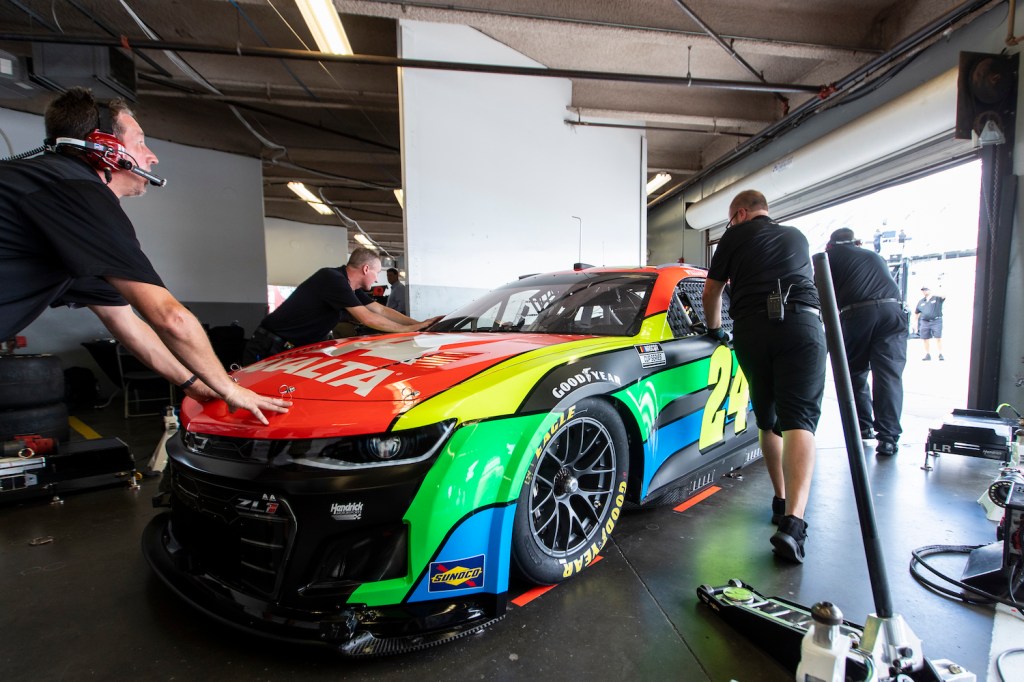 Four crew members roll the Next Gen Chevy race car out of a garage and onto a NASCAR track.
