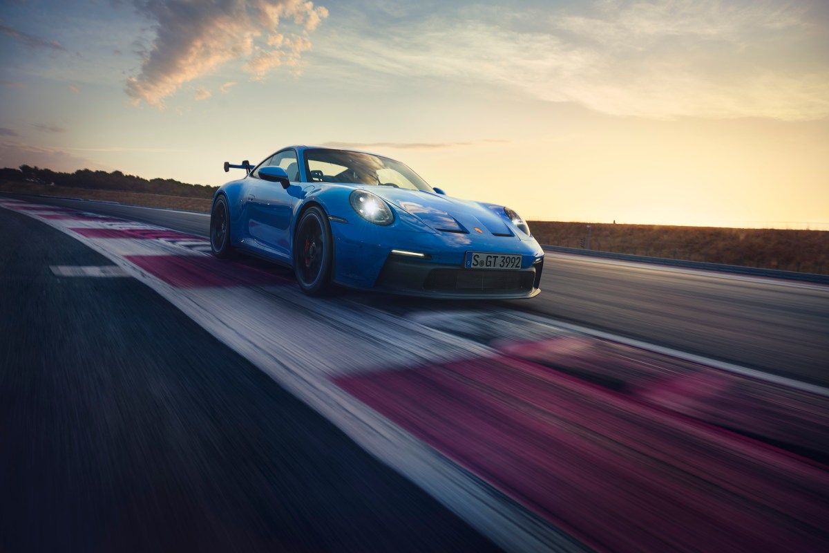 A 3/4 front view of a blue 2022 Porsche 911 GT3 cornering on a race track with a sunset in the background.