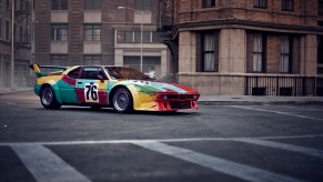 A 3/4 profile view of the Andy Warhol painted 1979 BMW m1 race car. Shown parked on a street with stone buildings in the background.