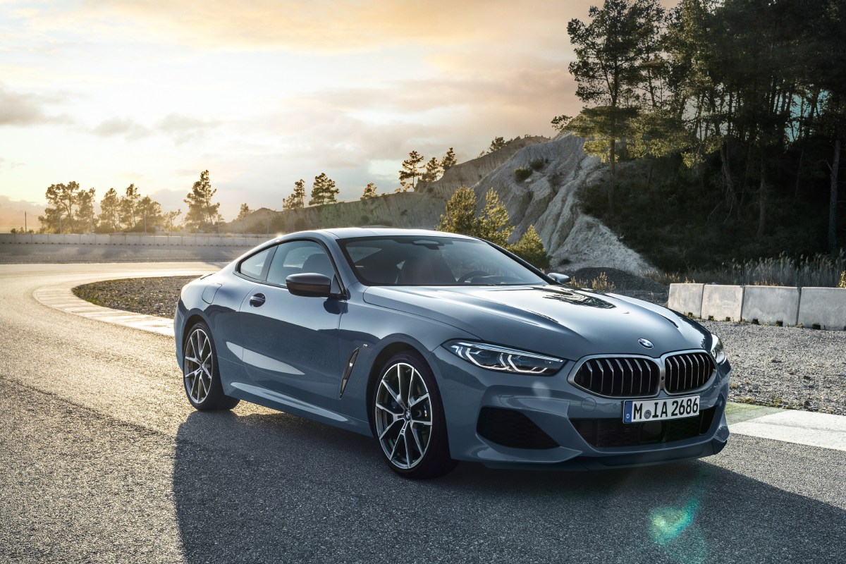 A 3/4 front view of a blue/grey 2022 BMW 8 Series Coupe parked at a corner of a race track. The background shows trees and rock cliffs.