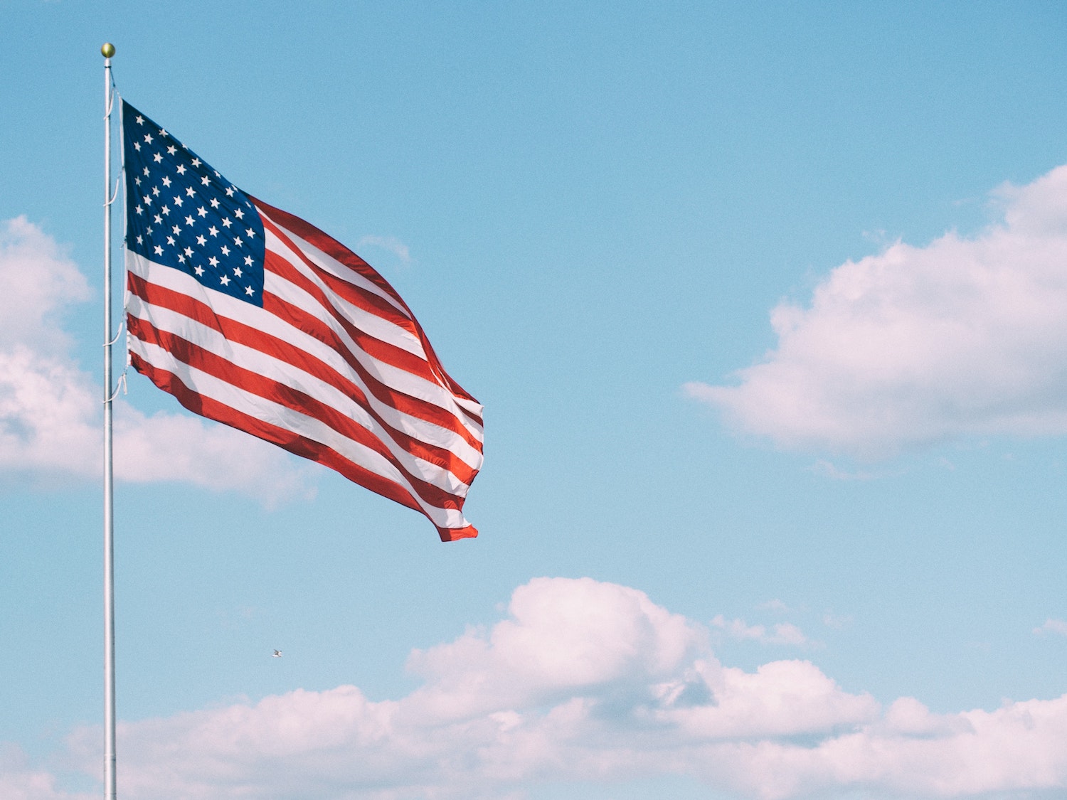 A large American flag blowing in the breeze in front of a light blue sky and fluffy white clouds.