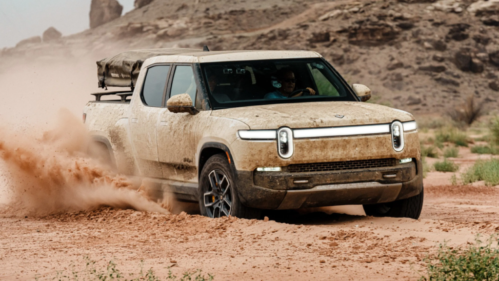 The Rivian R1T electric pickup truck covered in sand and dust as it drives through a field of dirt