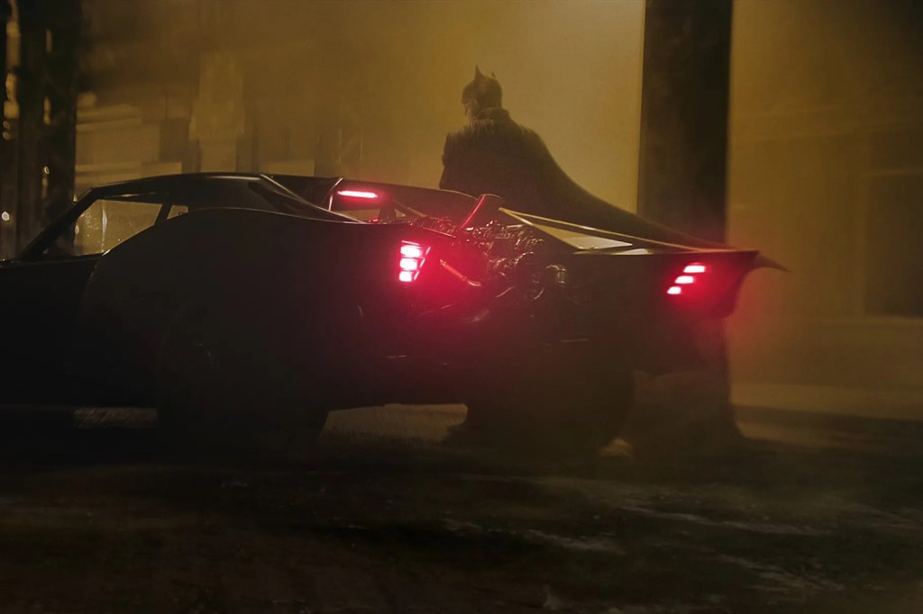 Robert Pattinson as batman next to his new muscle car batmobile parked on a dark, foggy street.