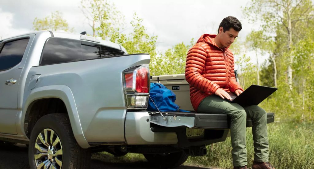 A man is sitting on the liftgate of  a gray Toyota Tacoma midsize truck while using a laptop. 