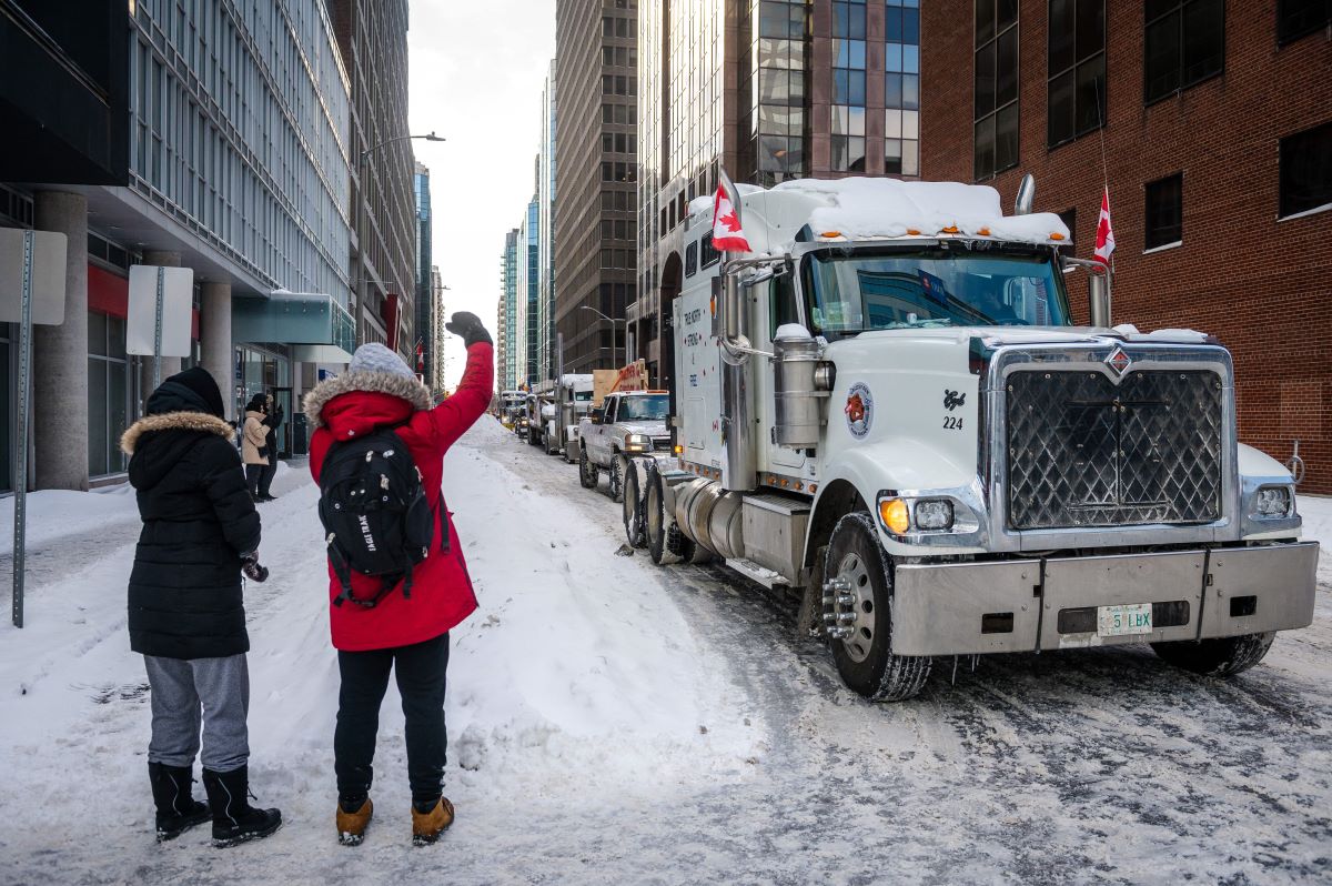 Semi-truck driving through snow at the trucker protests with people standing to the side.
