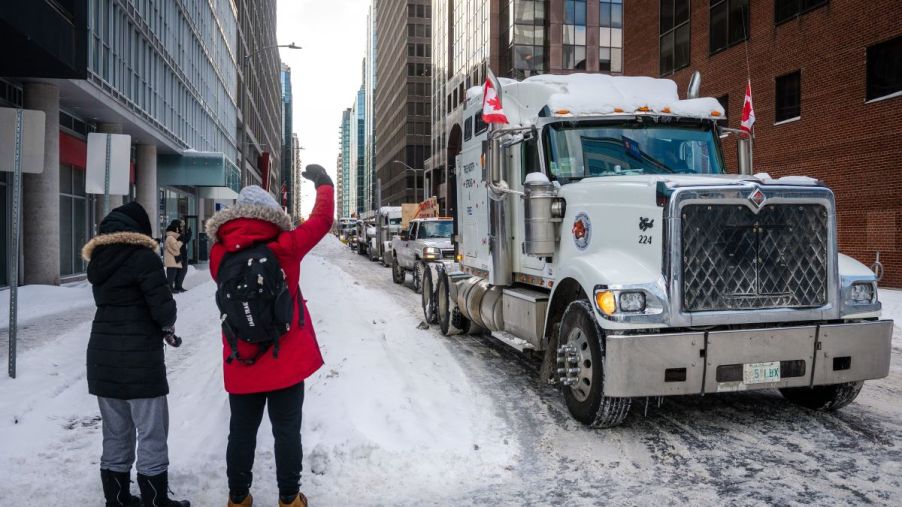 Semi-truck driving through snow at the trucker protests with people standing to the side.