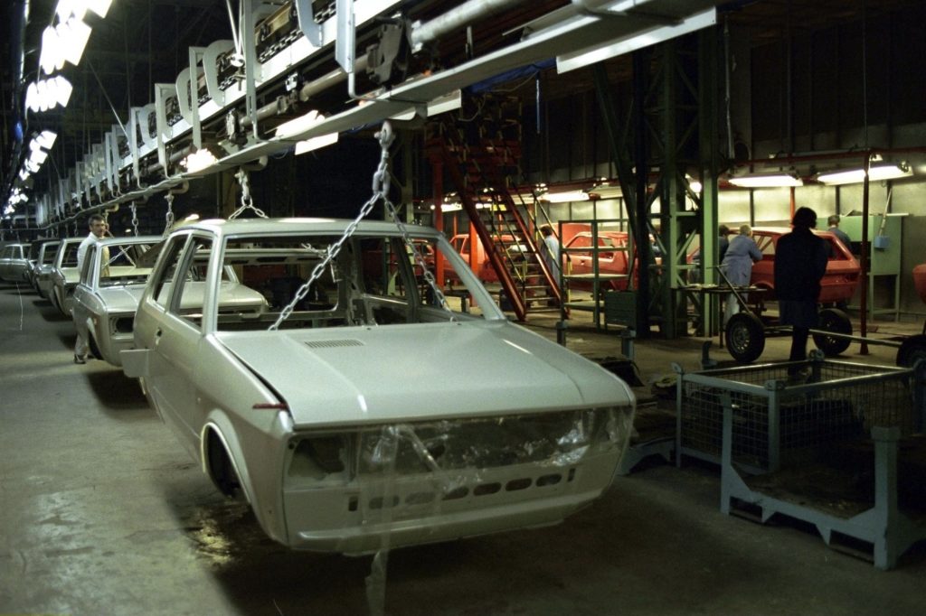 A white Yugo hatchback in a factory environment stripped of various parts. 