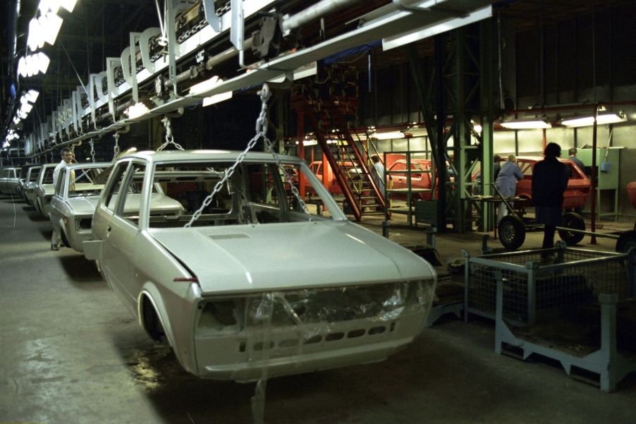 A white Yugo hatchback in a factory environment stripped of various parts.