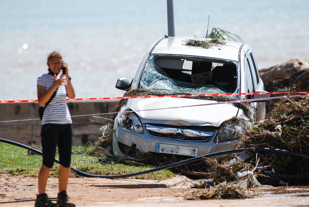 A woman speaking on the phone in front of a car damaged by floods on a street filled with mud and debris the day after flash floods.