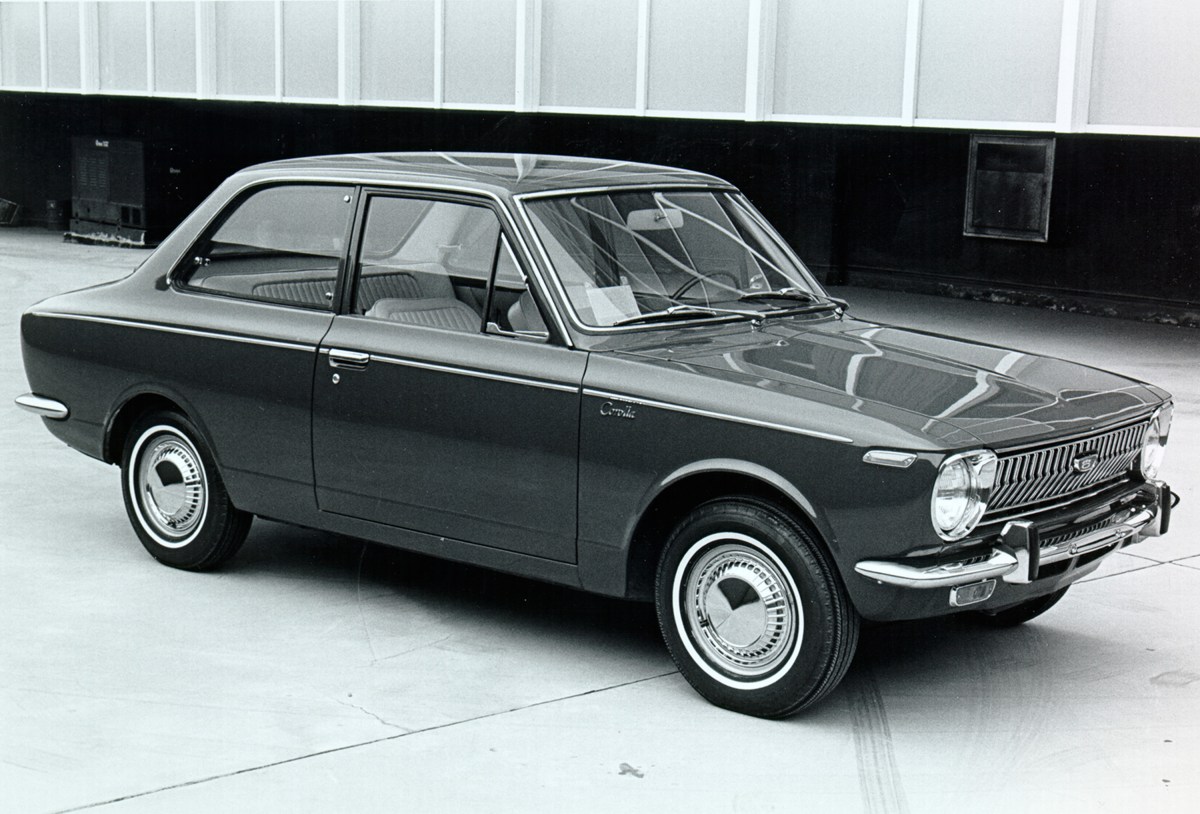 A black and white image of a 1st generation two-door Toyota Corolla parked in front of a building.